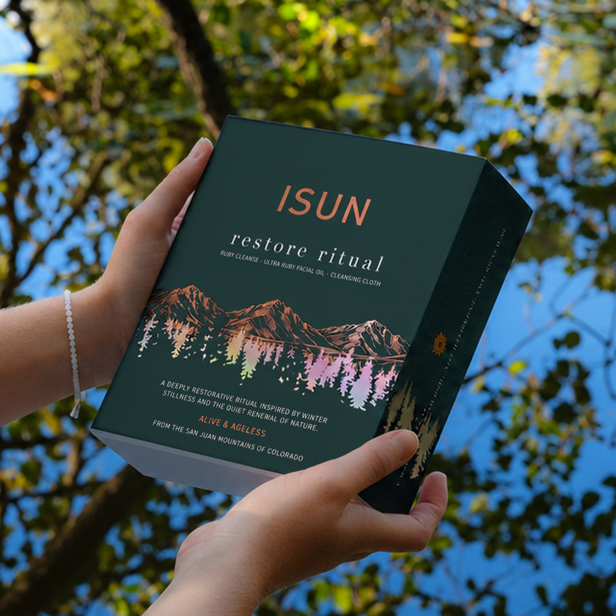 Hands holding an ISUN Restore Ritual gift box beside water reflecting green foliage.