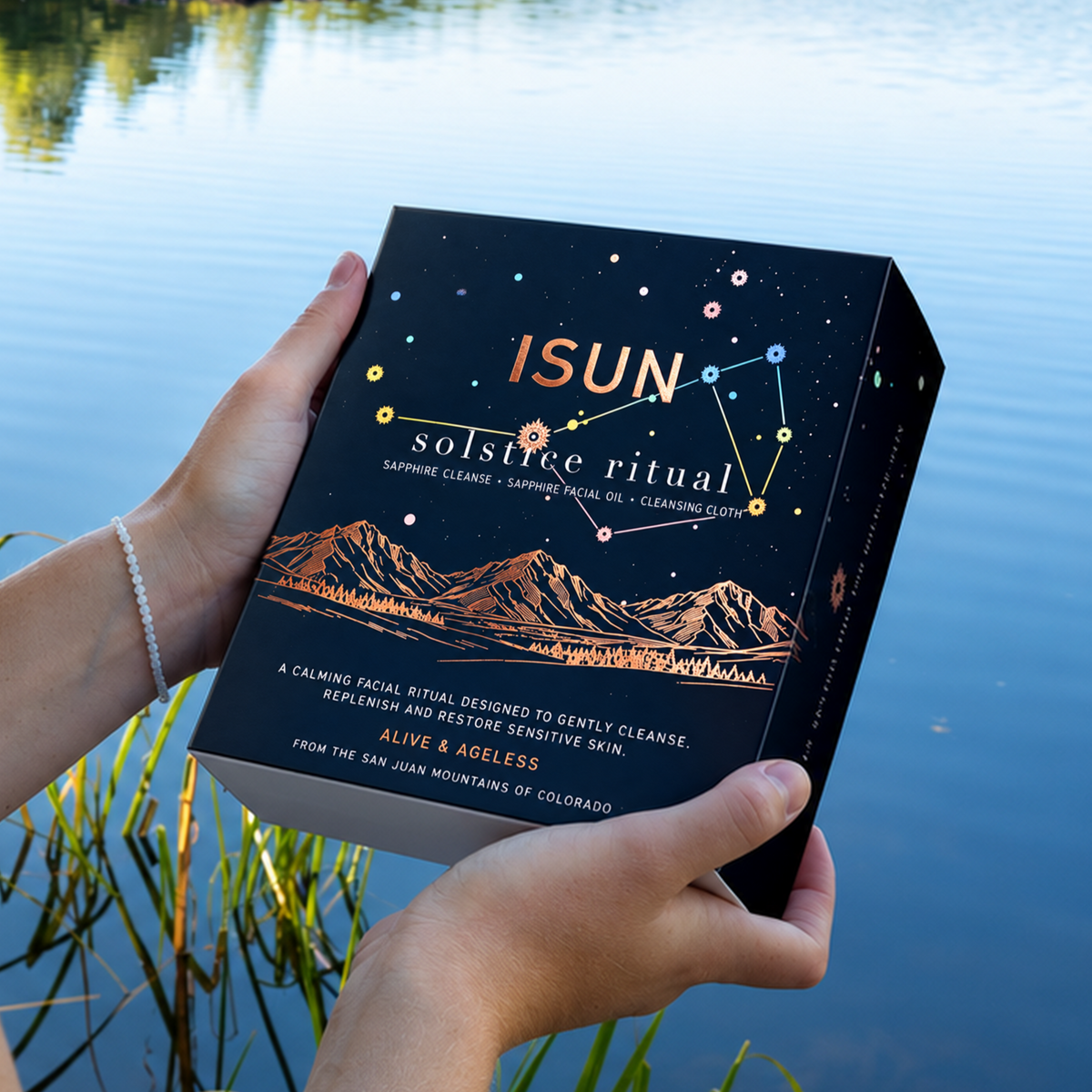 Hands holding an ISUN Solstice Ritual gift box with stars and a mountain illustration beside a lake.