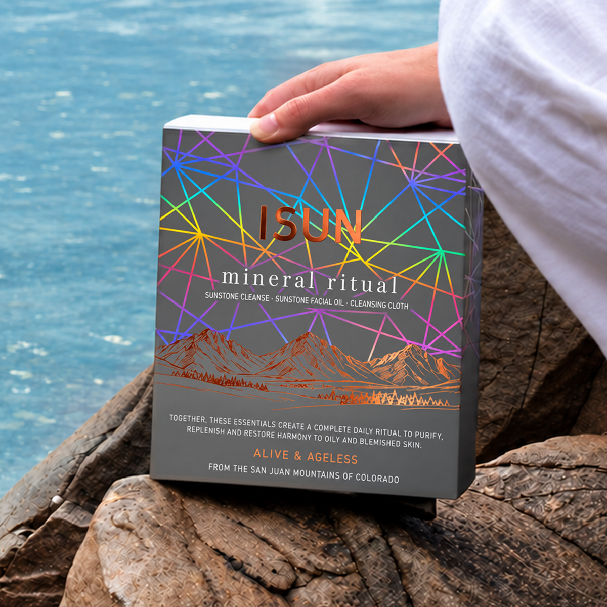 A person holding an ISUN Mineral Ritual gift box with geometric line art on rocks beside the water.