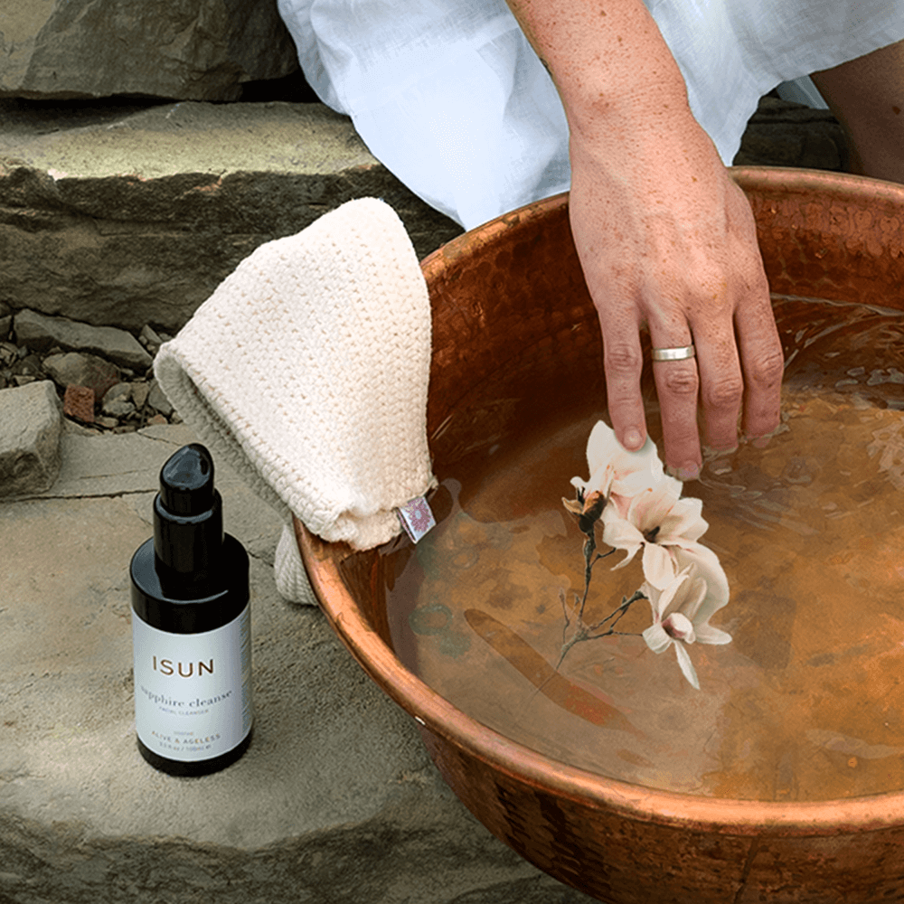 Person dipping a flower into a copper basin with a bottle of ISUN skincare product nearby.