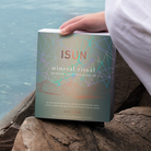 A person holding an ISUN Mineral Ritual gift box with geometric line art on rocks beside the water.