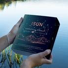 Hands holding an ISUN Solstice Ritual gift box with stars and a mountain illustration beside a lake.